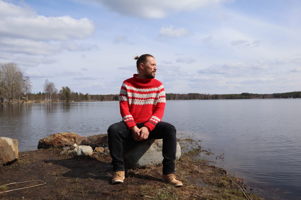 Panu Huotari istuu nuotiopaikan kivellä rantamaisemassa.